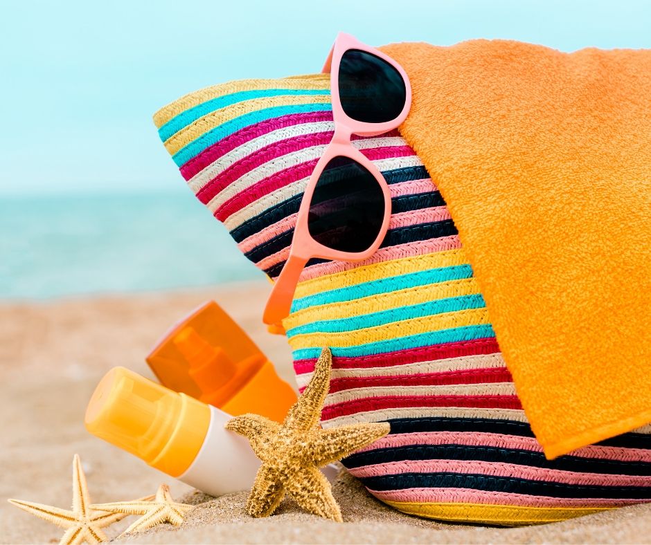 beach bag sitting on sand, with sunglasses and sunscreen